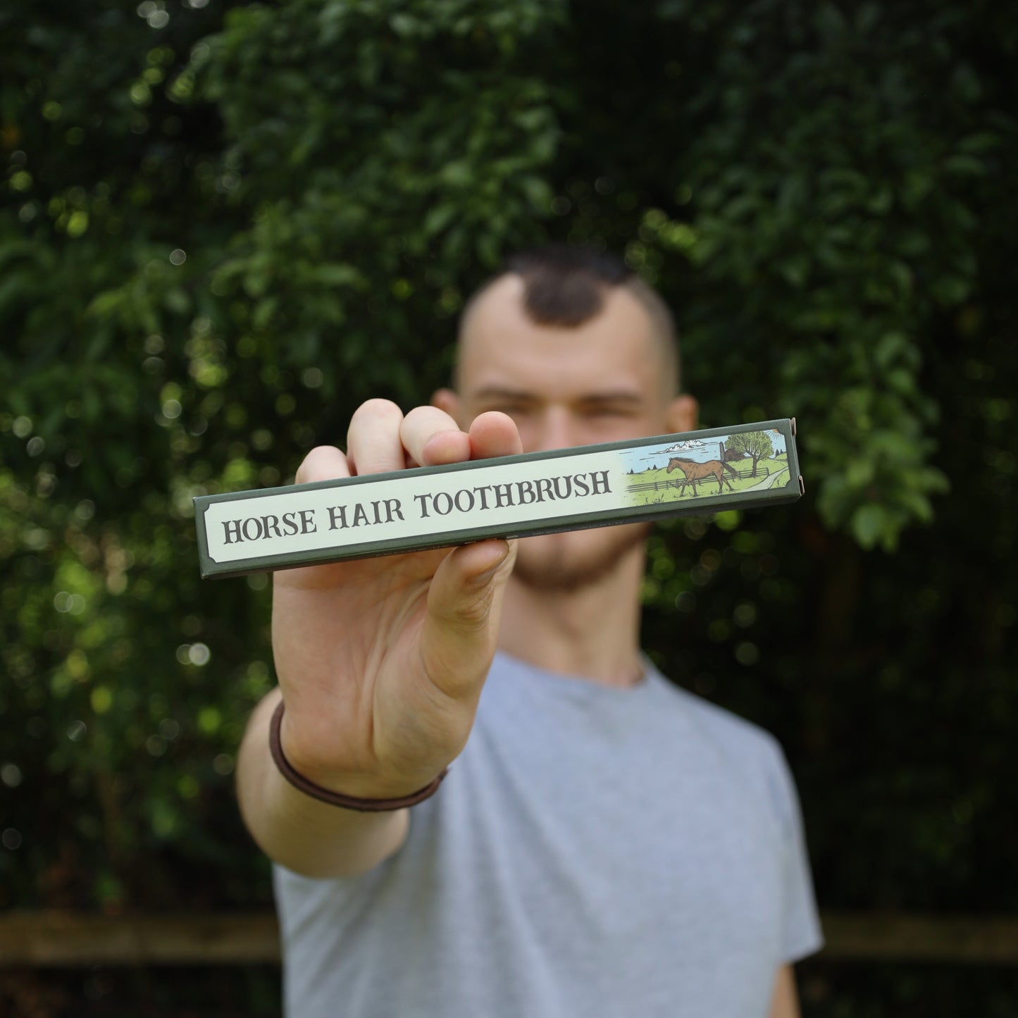 Portrait-style image of a man holding up the ESO World toothbrush box toward the camera, centered in natural greenery. The model’s face is slightly blurred, drawing focus to the product and its label. This lifestyle shot adds a human element to the product listing, increasing emotional relatability. Optimized for keywords like “sustainable gift ideas,” “bamboo toothbrush lifestyle photo,” “horsehair toothbrush held by person,” or “natural oral care with traditional materials.” Useful for retailers, blogs, o